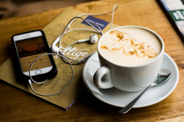 Latte, iPhone with podcast app showing, white earbuds with cables on top of a menu. Everything is on a light wood tabletop.