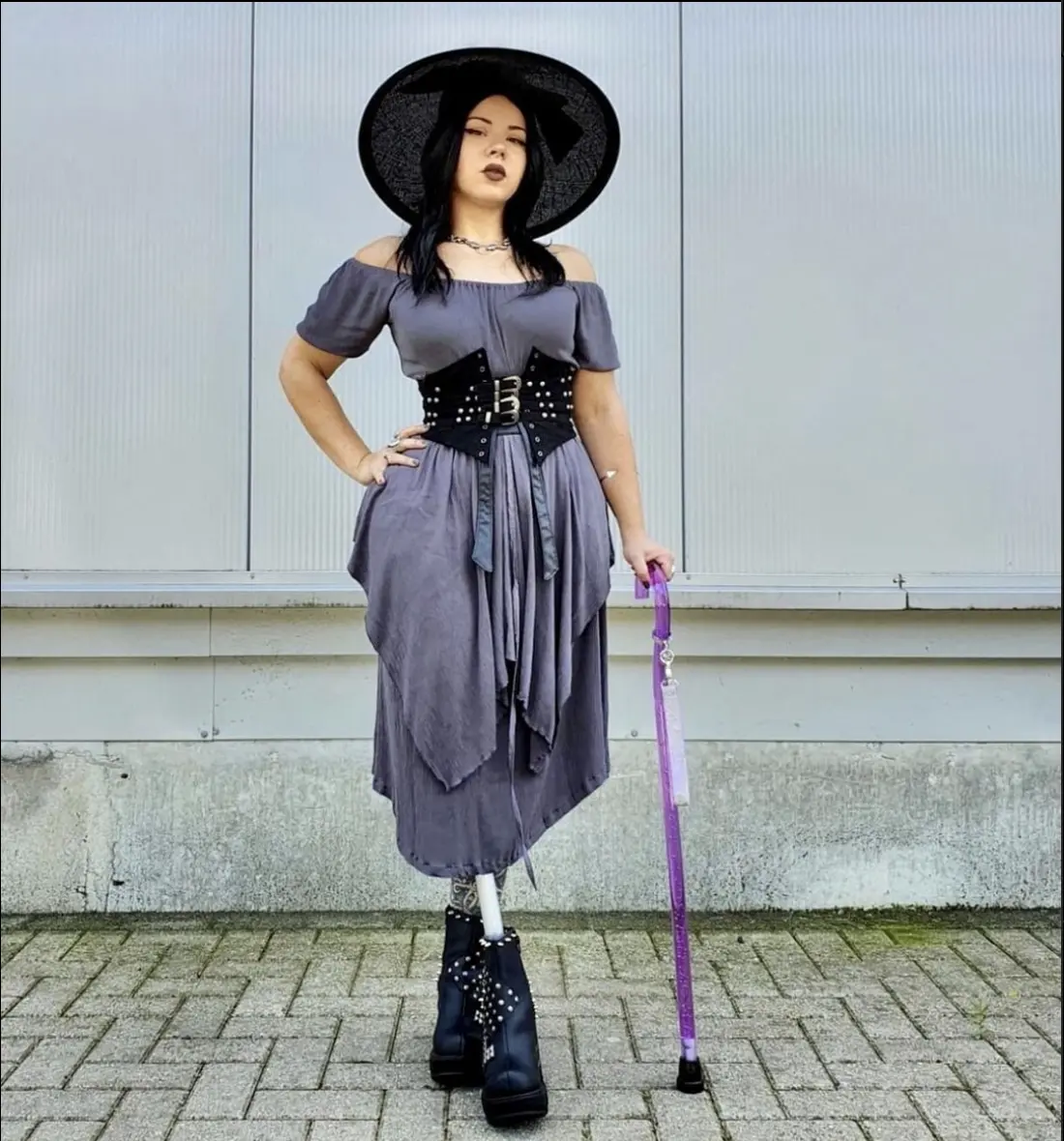 'Young woman wearing a high fashion, light greyish purple off the shoulder short sleeved dress with a layered asymetical bottom hem. She has on a very wide brimbed matching hat. She has straight black shoulder length hair, and appears to be Asian. She has a translucent bright purple curved handle walking cane with bubbles inside it and a light pink ribbon strap hanging from the handle. She's also wearing a very wide leather black goth type belt and platform boots. One leg has tatoos and the other might be a prostetic'
