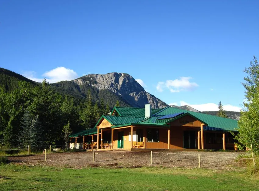 Photo of a wood log lodge with a green roof in the mountains, There’s a Canadian flag flying on a flagpole in the middle.